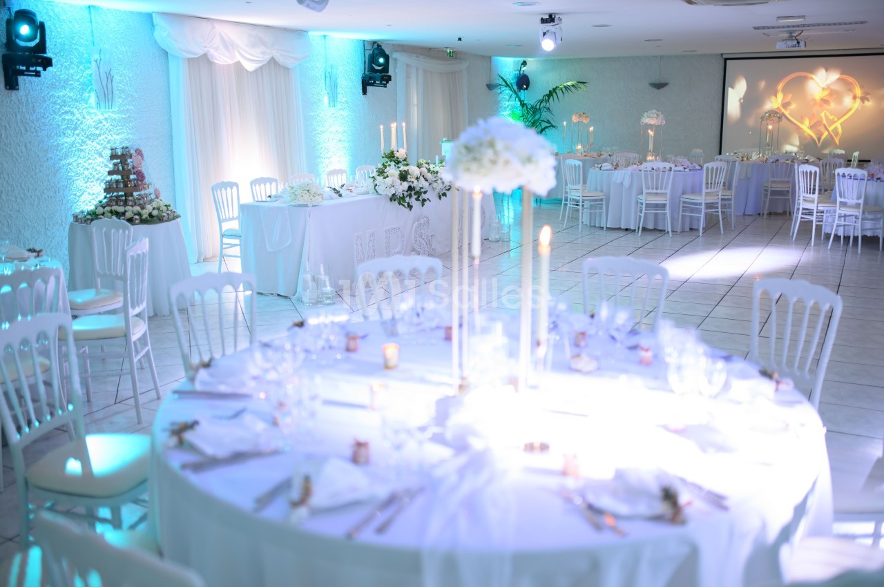 Salle de réception décorée avec des tables rondes, nappes blanches, chandeliers et éclairage bleu tamisé.