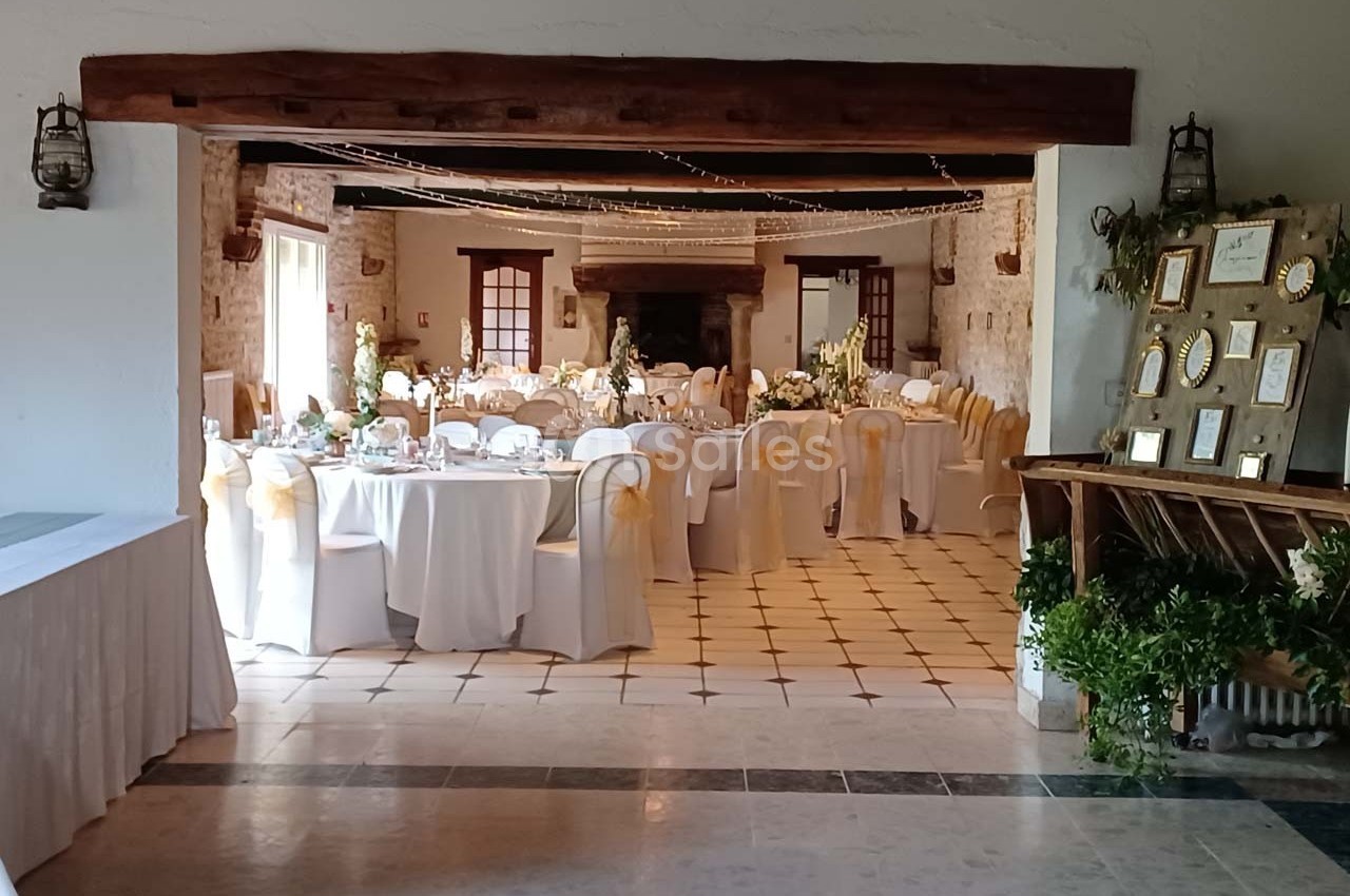 Salle de réception décorée avec des tables rondes, nappes blanches, chaises ornées de rubans dorés et cadres exposés.