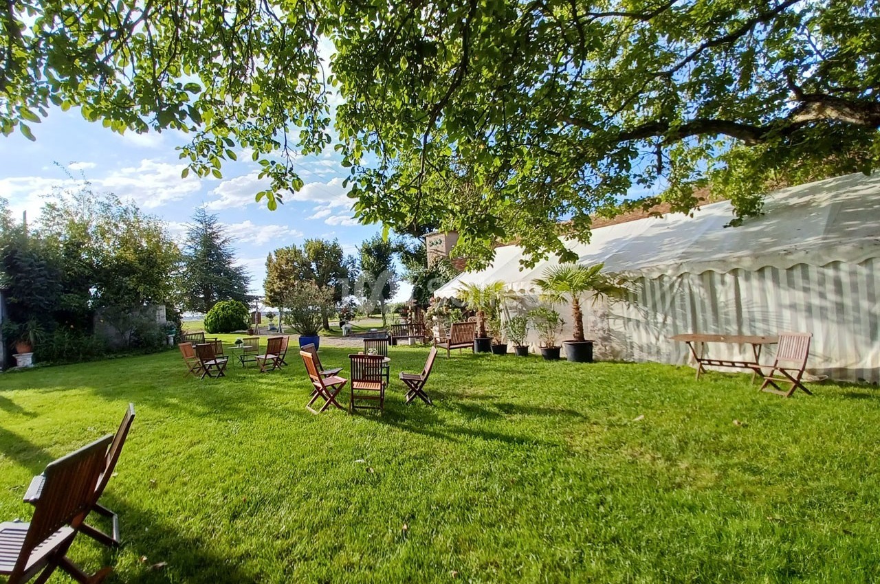Chaises et tables en bois disposées sur une pelouse verdoyante, sous des arbres, près d'une grande tente blanche.