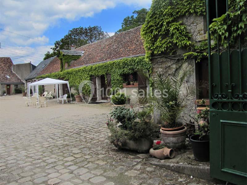 Cour pavée avec bâtiments en pierre couverts de lierre, tente blanche et pots de fleurs disposés devant.