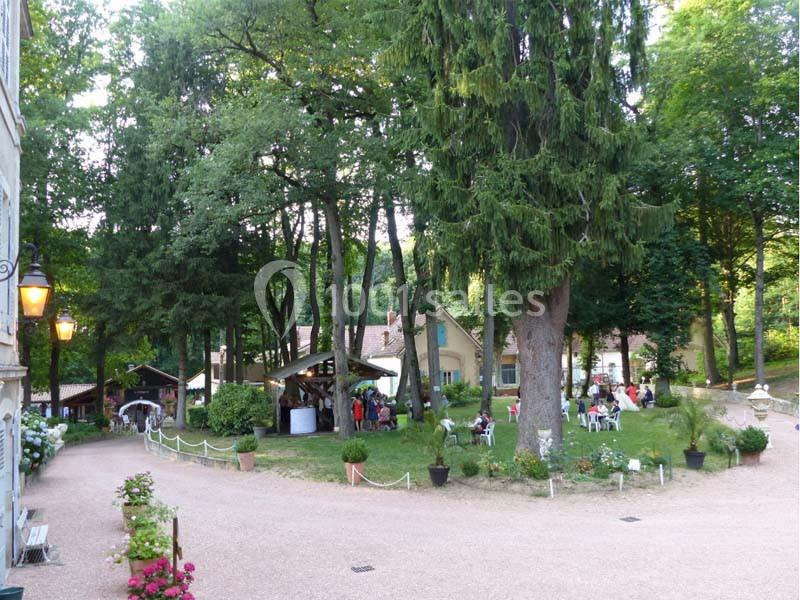 Jardin arboré avec des tables et des chaises occupées, entouré de bâtiments et éclairé par des lampes en soirée.