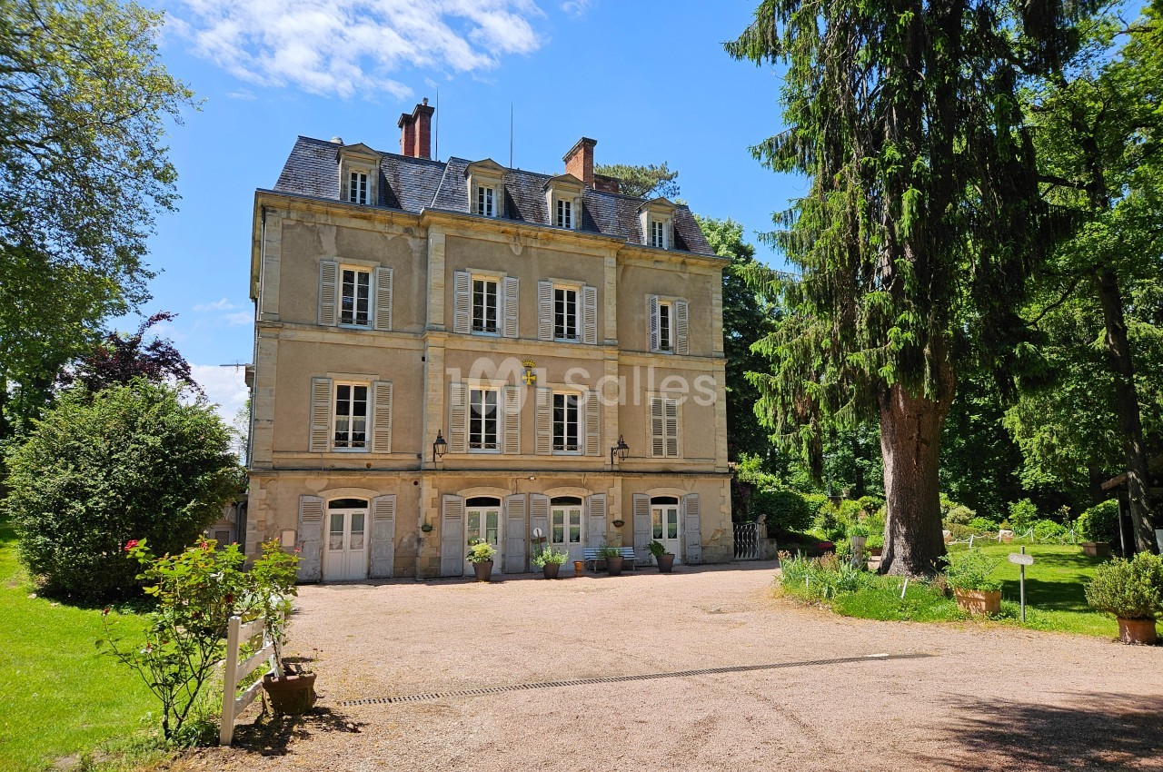Façade d'un manoir en pierre entouré d'arbres et d'un jardin, avec une allée gravillonnée au premier plan.