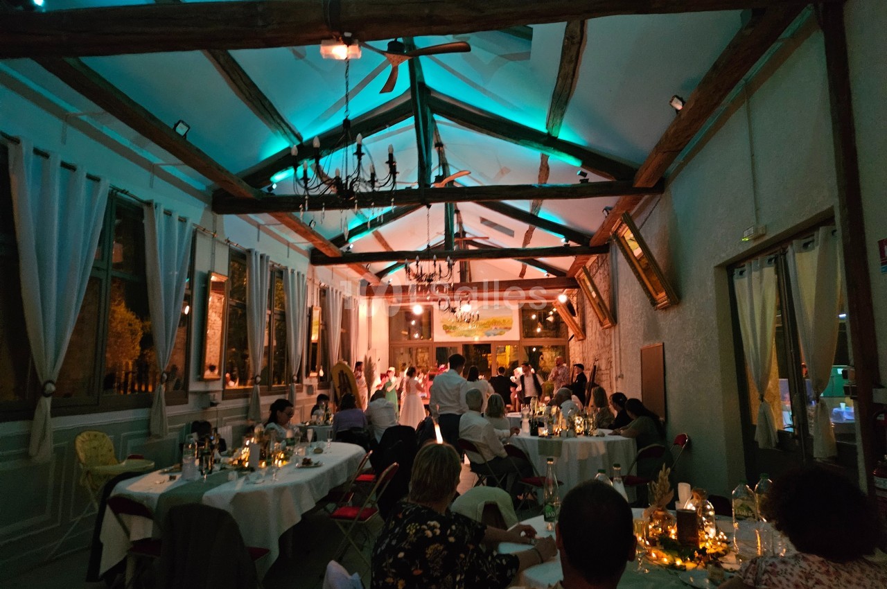 Salle de réception éclairée en soirée, avec des invités assis à des tables décorées et une piste de danse au fond.