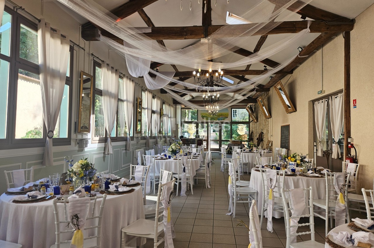 Salle de réception lumineuse décorée pour un événement, avec tables rondes dressées et voilages suspendus au plafond.