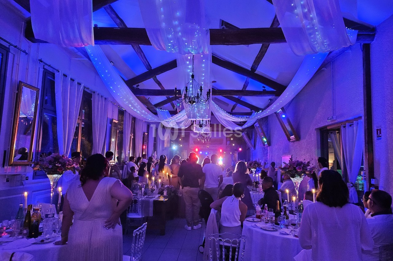 Salle de réception décorée avec des guirlandes lumineuses, des invités assis et debout lors d'une soirée festive.