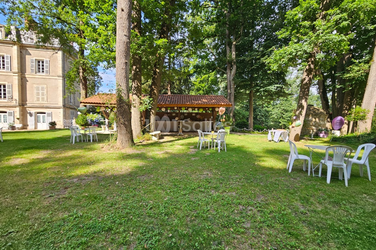 Espace extérieur avec pelouse, tables et chaises blanches, cabanon décoré et arbres entourant un bâtiment ancien.