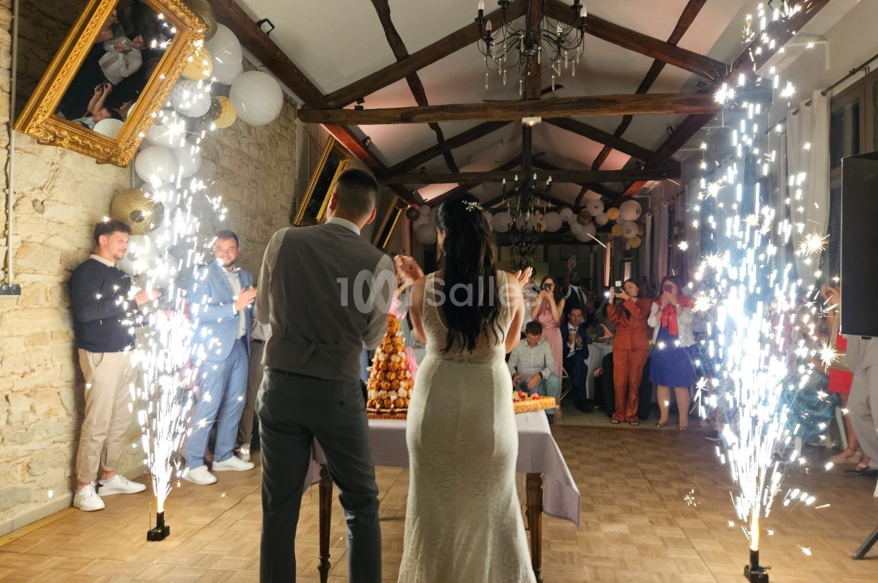 Un couple de mariés coupe une pièce montée dans une salle décorée, entouré d'invités et de fontaines lumineuses.
