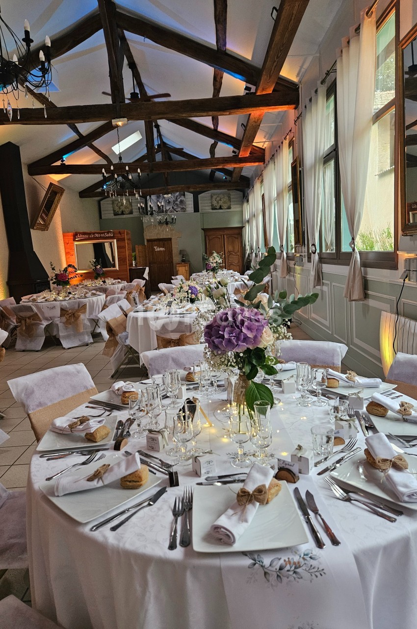 Salle de réception décorée avec des tables rondes élégamment dressées, ornées de fleurs et éclairées par une lumière…