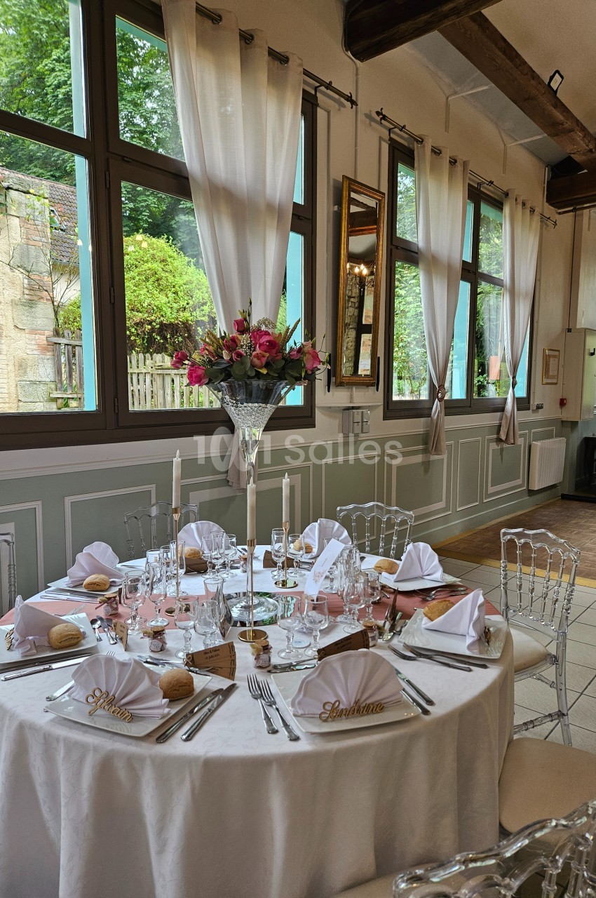 Table élégamment dressée avec nappes blanches, vaisselle raffinée, bougies et bouquet de fleurs, dans une salle lumineuse.