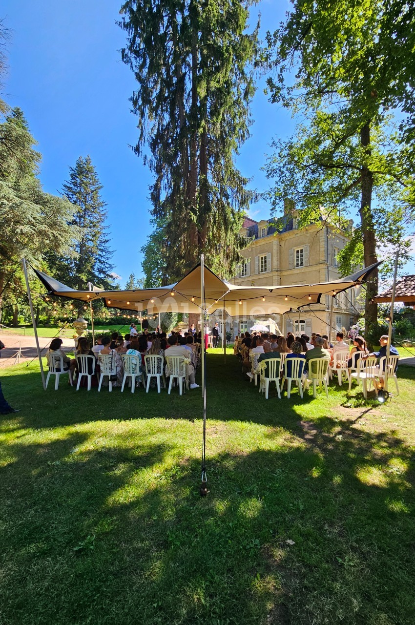 Cérémonie en plein air sous une tente, avec des invités assis sur des chaises blanches, devant un bâtiment ancien.