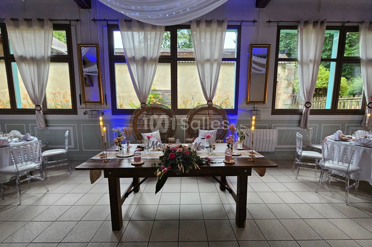 Salle de réception décorée avec une table en bois ornée de fleurs, deux fauteuils et des bougies devant des fenêtres.