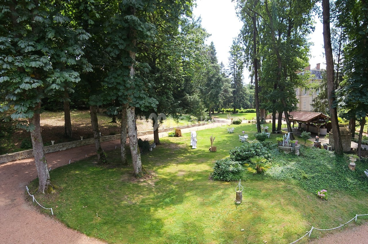 Jardin verdoyant avec pelouse, arbres, allée en gravier et quelques bancs près d'un bâtiment en arrière-plan.