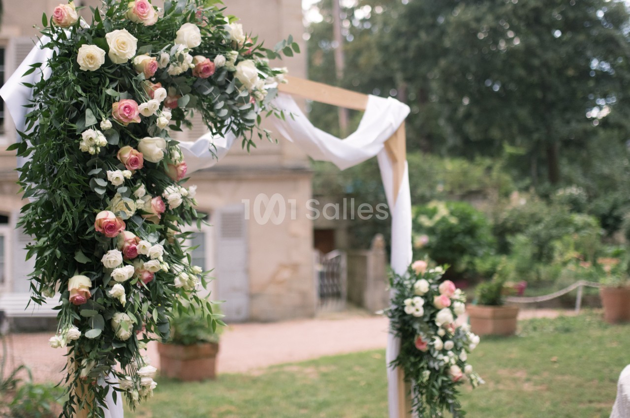 Arche de mariage en bois décorée de fleurs blanches et roses, entourée d'un jardin verdoyant.