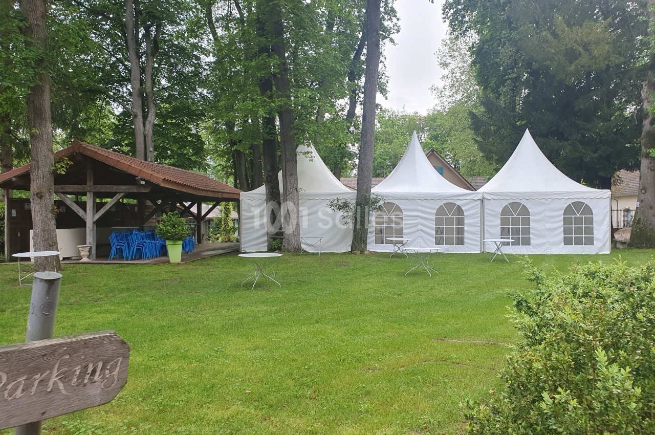 Tentes blanches et abri en bois installés dans un espace vert entouré d'arbres, avec des tables disposées sur la pelouse.