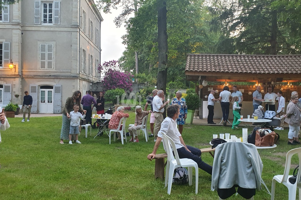 Groupe de personnes rassemblées dans un jardin, près d'un bâtiment ancien et d'un espace couvert avec un bar.