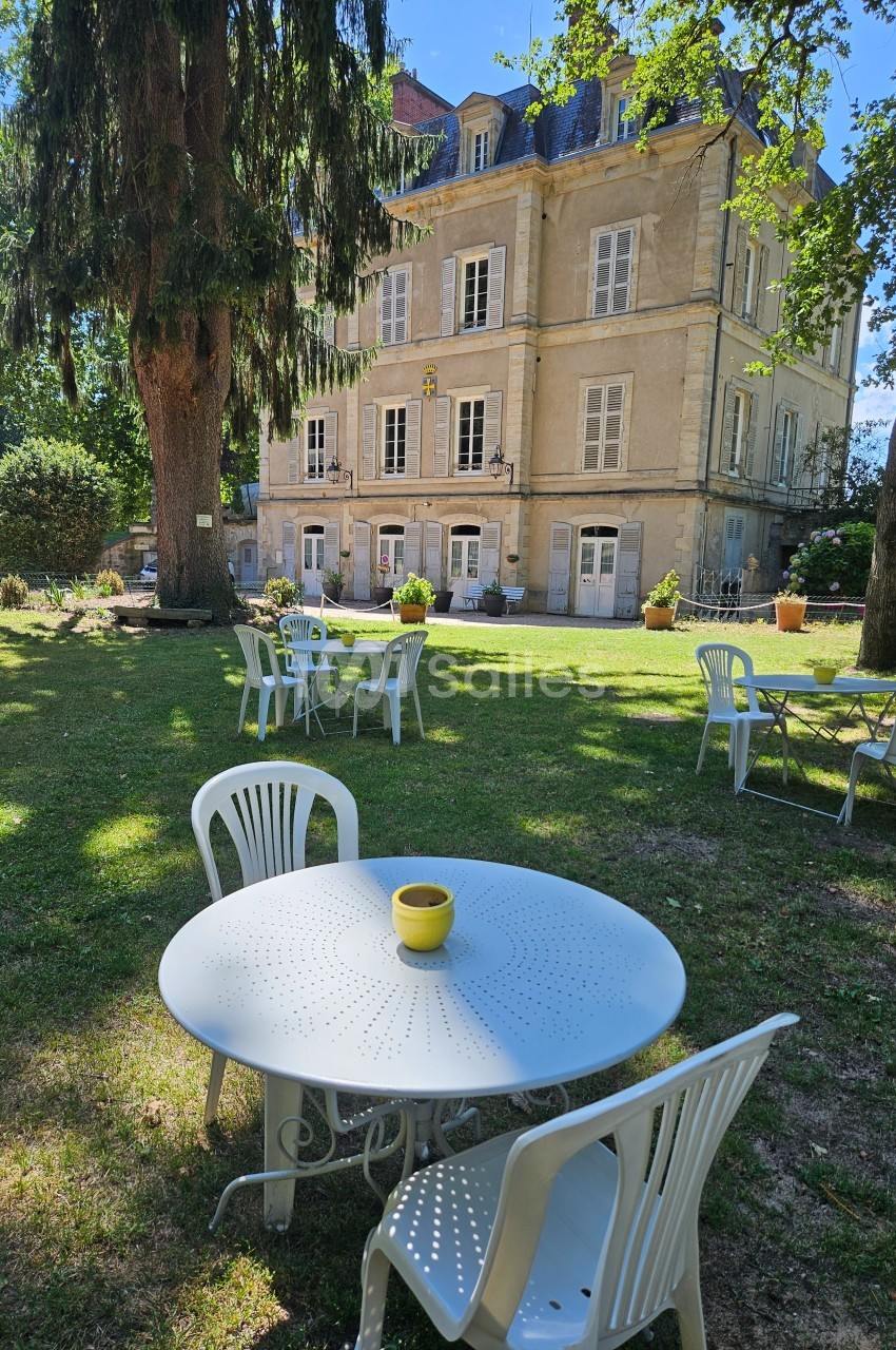 Tables et chaises blanches disposées dans un jardin ombragé devant un grand bâtiment ancien en pierre.