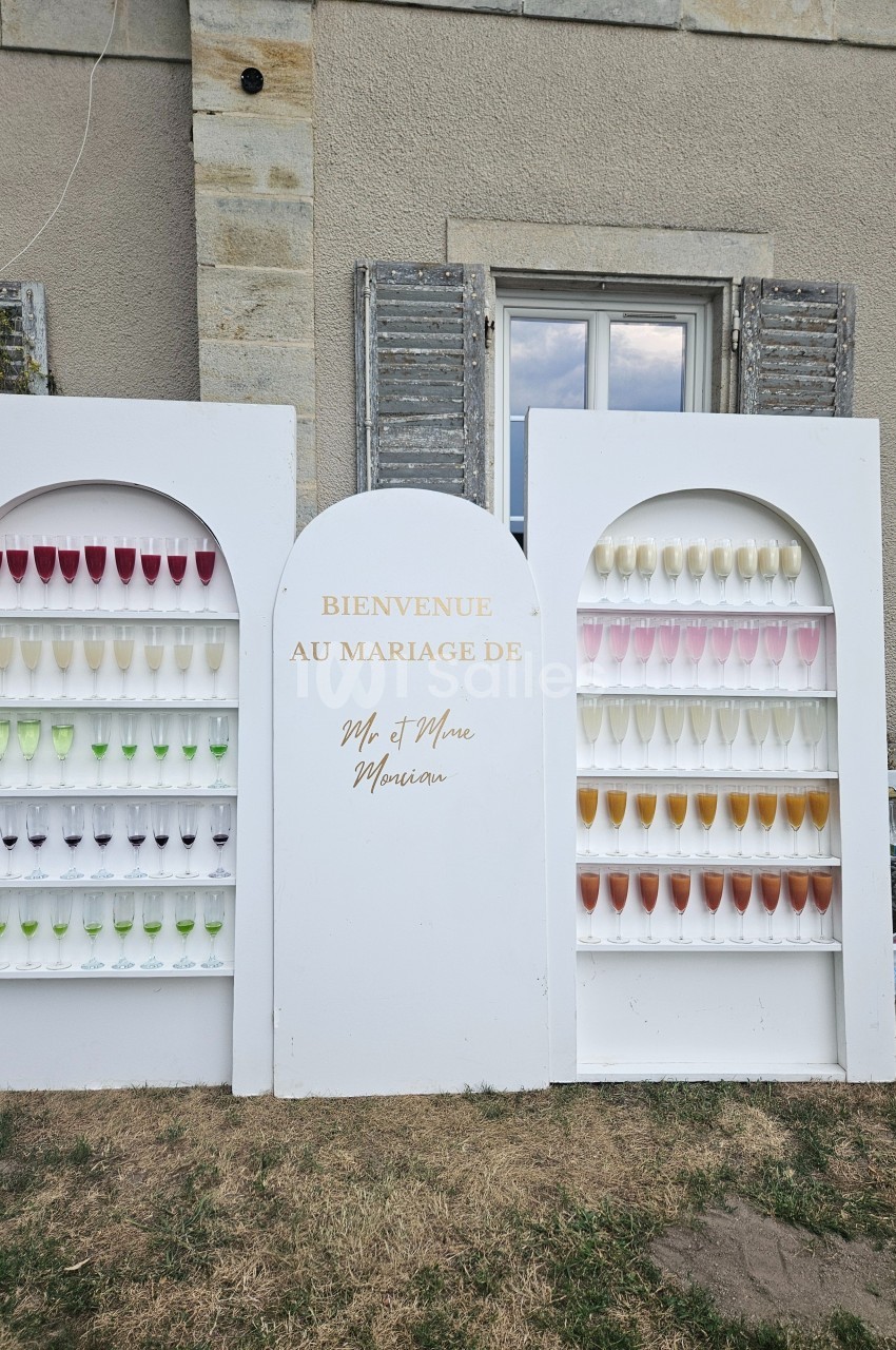 Mur décoratif avec des rangées de coupes de champagne colorées, installé devant un bâtiment en pierre.