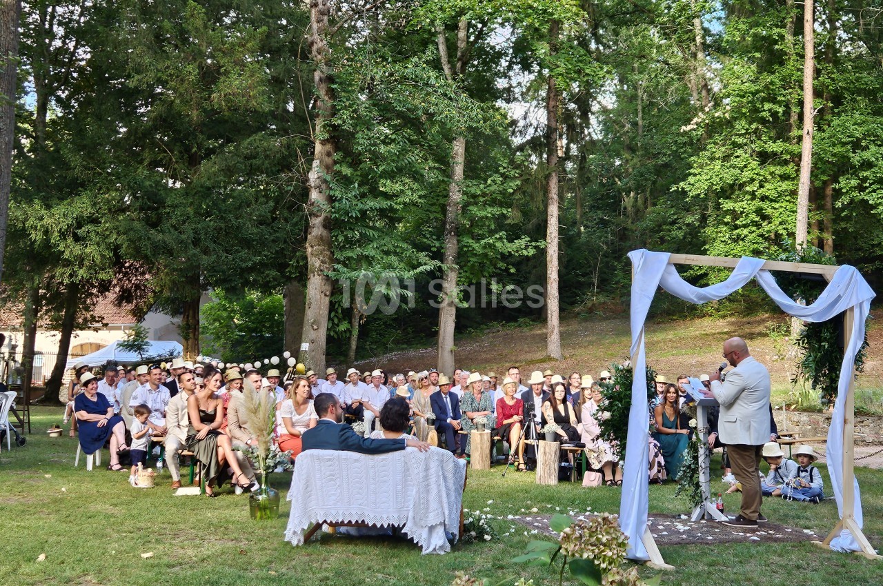 Cérémonie en plein air dans un jardin boisé, avec des invités assis et un officiant devant une arche décorée.