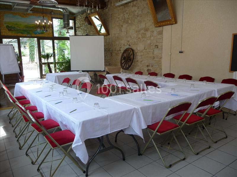 Salle de réunion avec tables disposées en U, nappes blanches, chaises rouges et écran de projection en arrière-plan.