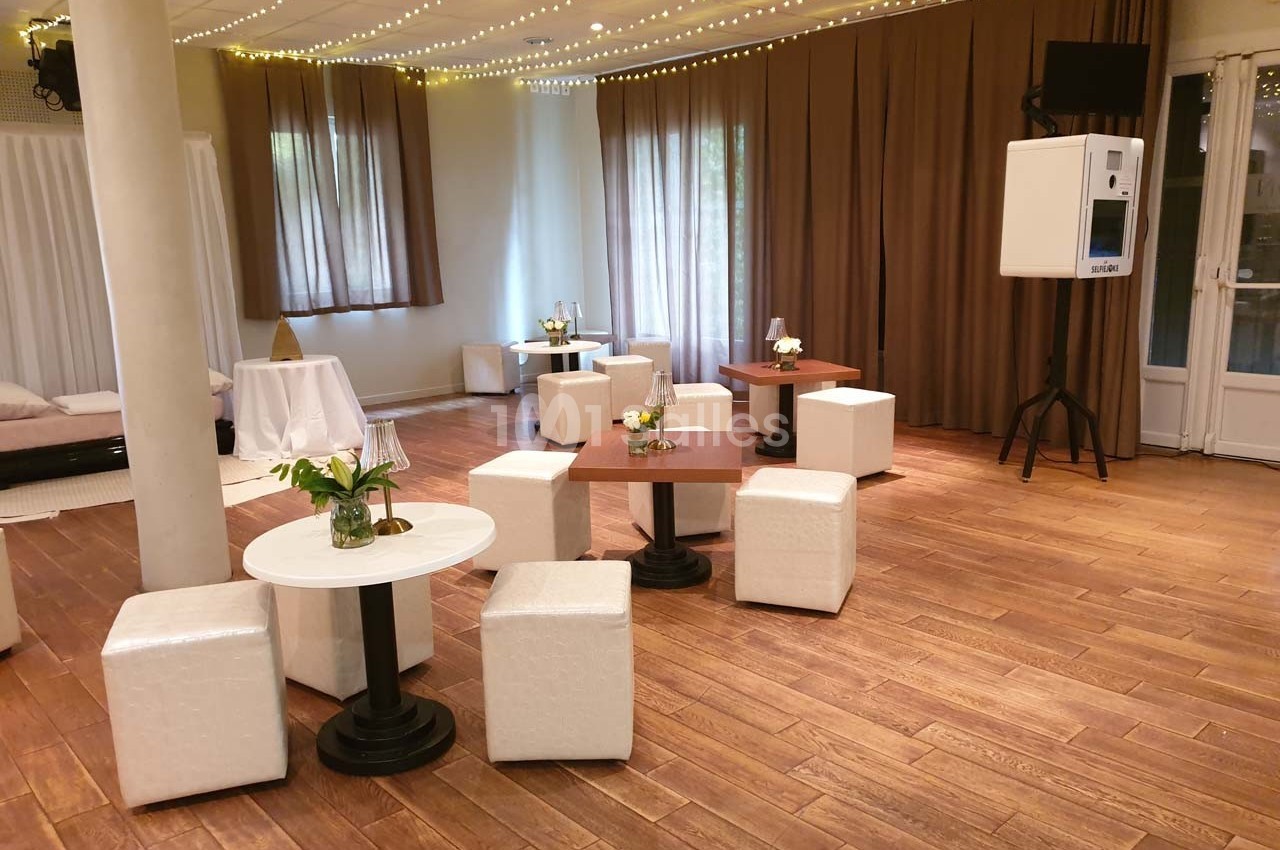 Salle lumineuse avec parquet, tables basses, poufs blancs, rideaux marron et guirlandes lumineuses au plafond.