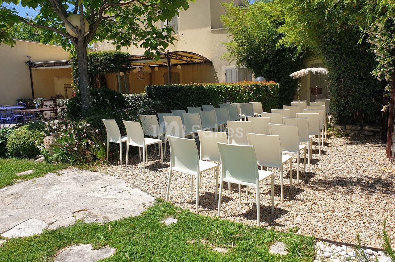 Chaises blanches disposées en rangées dans un jardin ensoleillé, entouré de verdure et de bâtiments clairs.
