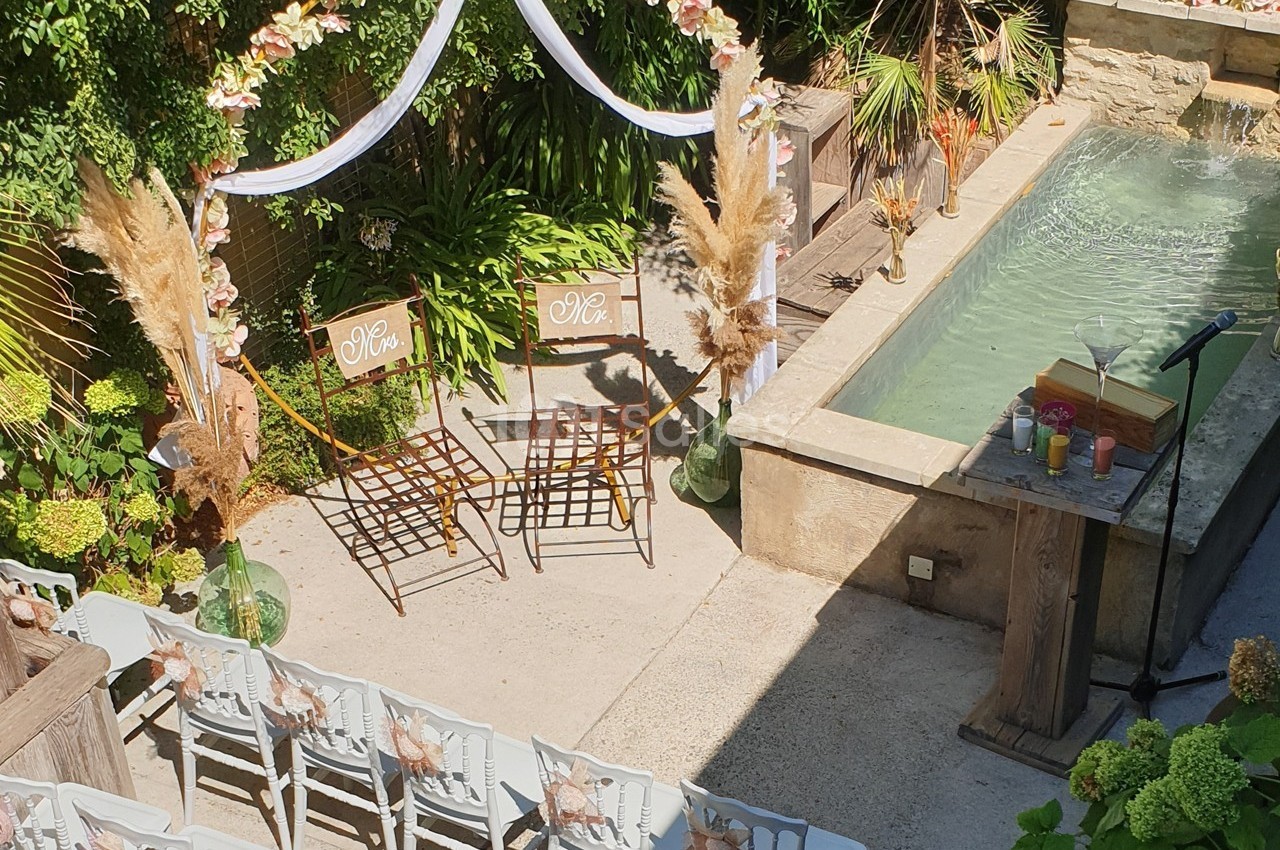 Chaises décorées sous une arche fleurie près d'un bassin, disposées pour une cérémonie en extérieur.