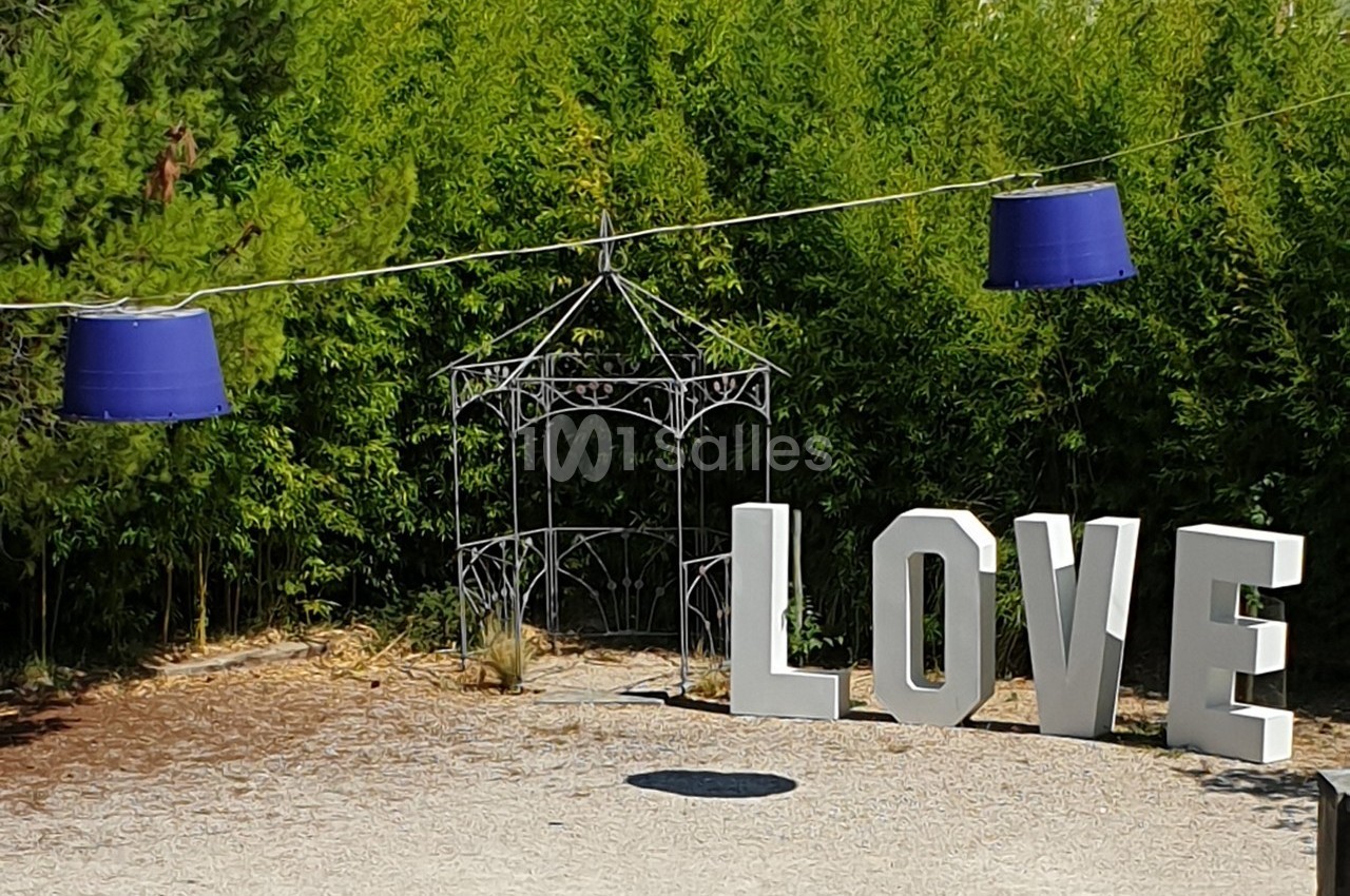 Lettres blanches formant le mot ’LOVE’ dans un jardin avec des bambous, une structure métallique et des lampes suspendues.