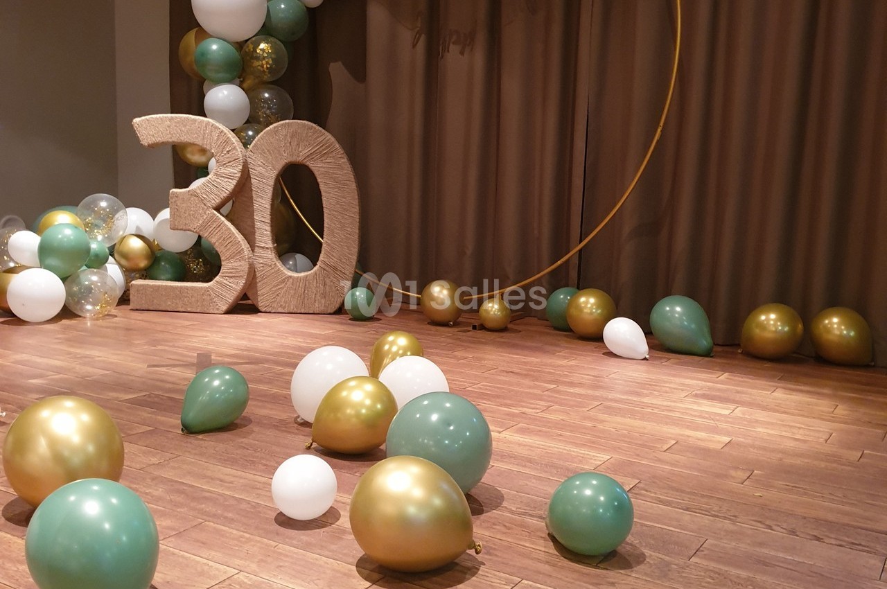 Décoration de fête avec ballons dorés, verts et blancs, chiffre 30 en relief et rideaux en arrière-plan.
