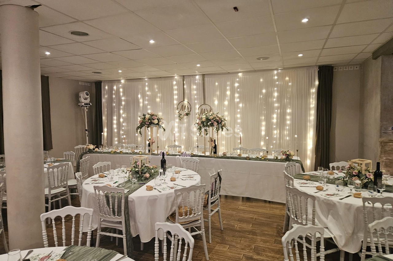 Salle de réception décorée pour un événement, avec tables rondes dressées et guirlandes lumineuses en arrière-plan.