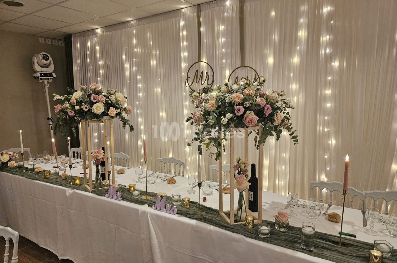 Table de réception décorée avec des fleurs, bougies et guirlandes lumineuses, dans une salle élégante.