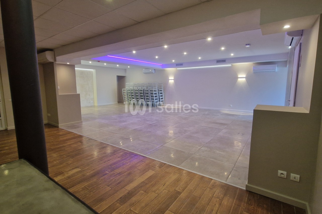 Salle vide avec parquet et dallage clair, éclairage encastré blanc et bandeau LED bleu au plafond, chaises empilées au fond.