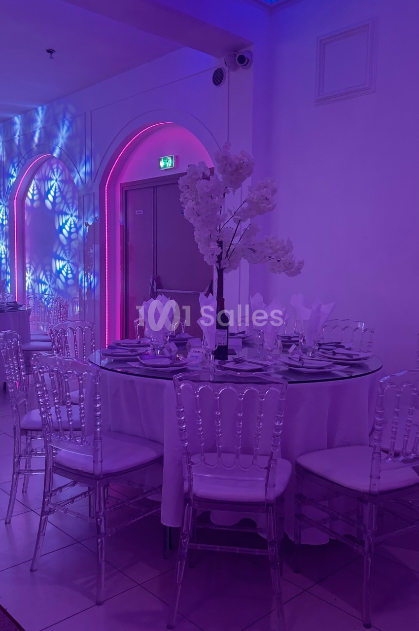 Salle de réception avec table ronde décorée, chaises transparentes et éclairage violet et bleu en arrière-plan.