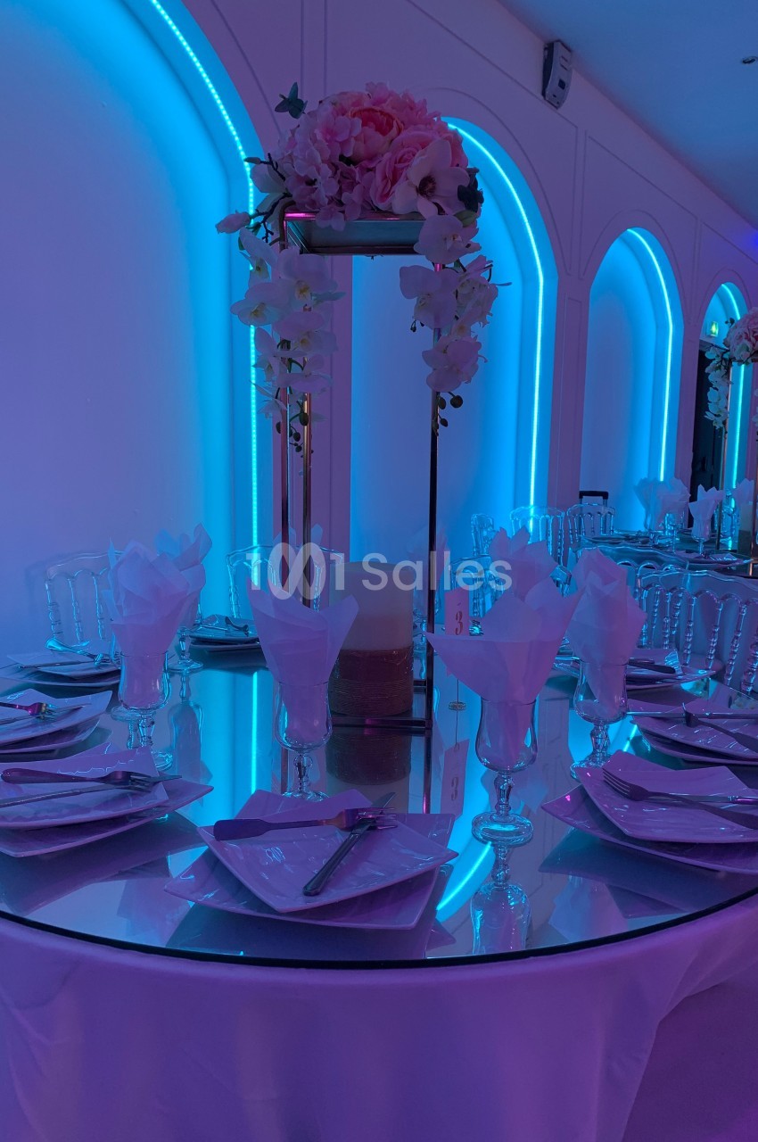 Table ronde décorée avec des assiettes, verres et serviettes, éclairée par une lumière bleue et ornée de fleurs.