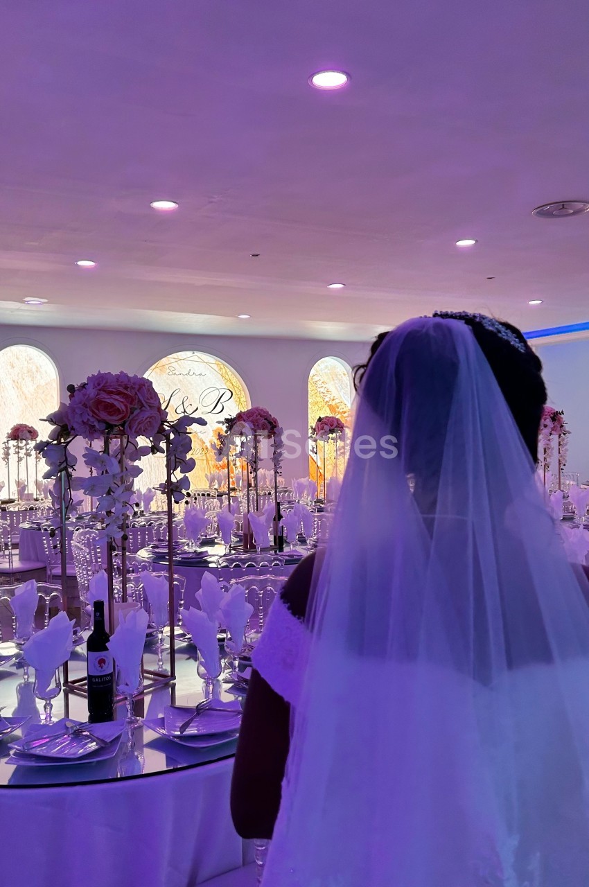 Une mariée de dos dans une salle de réception décorée avec des tables dressées et des arrangements floraux.