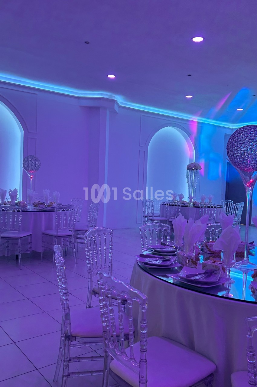 Salle de réception éclairée en violet avec tables dressées, chaises transparentes et décorations élégantes.