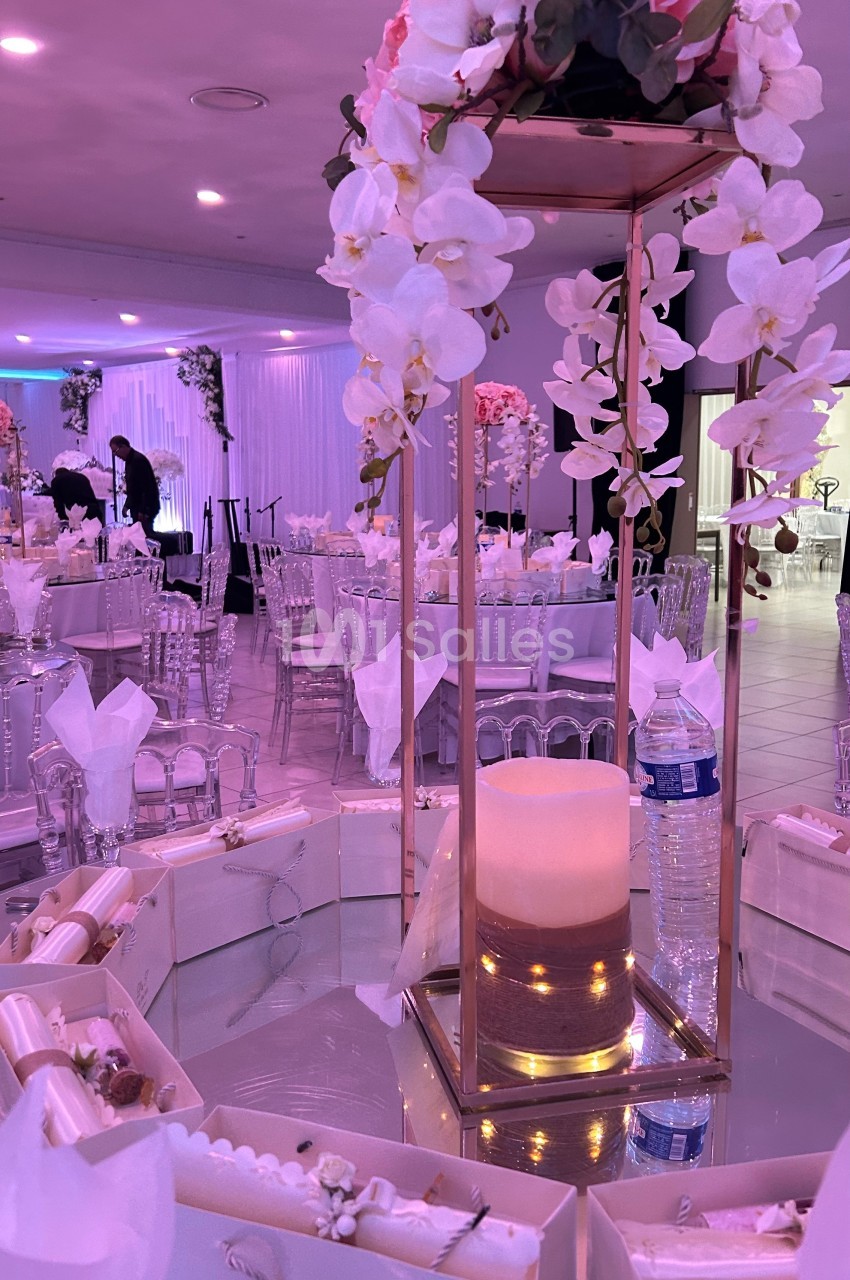 Centre de table décoratif avec bougie, fleurs blanches et roses, entouré de cadeaux sur une table de réception élégante.