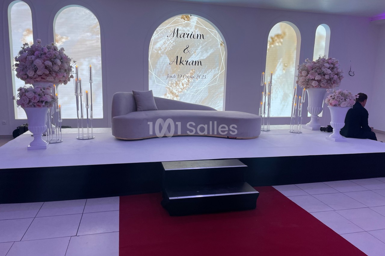 Décor de scène avec canapé beige, fleurs blanches et bougies, fond lumineux affichant des prénoms et une date.