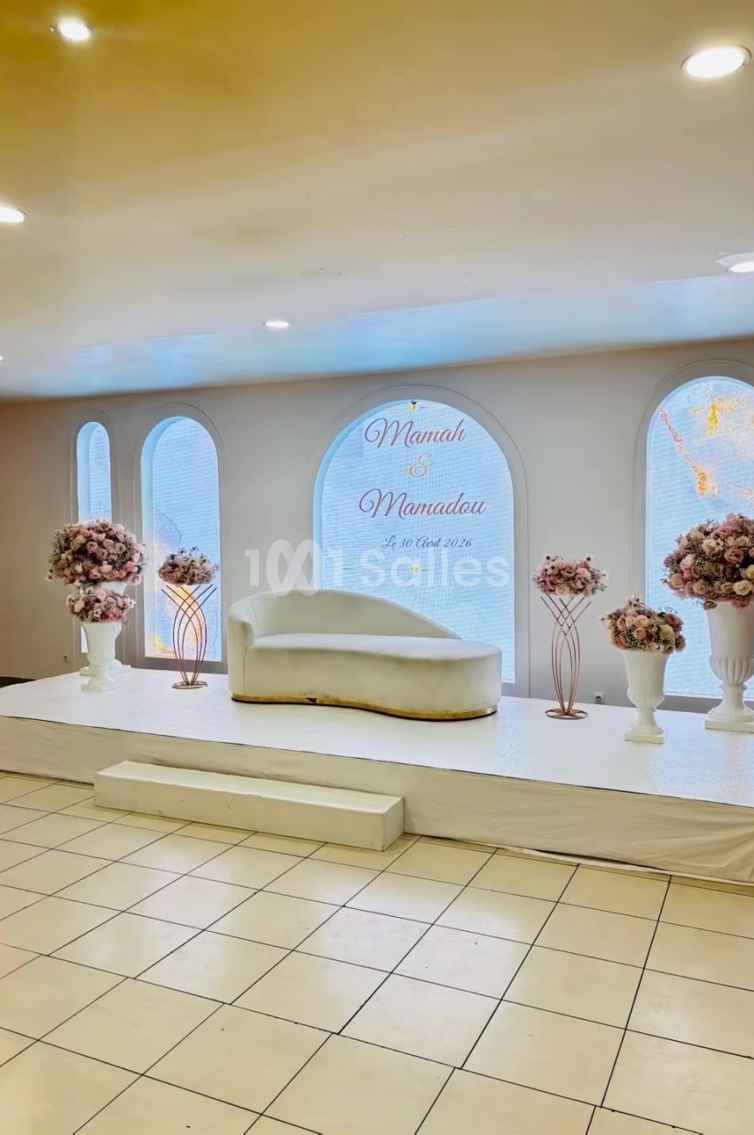 Décor de salle avec canapé blanc, fleurs roses sur piédestaux et inscription murale célébrant un mariage.