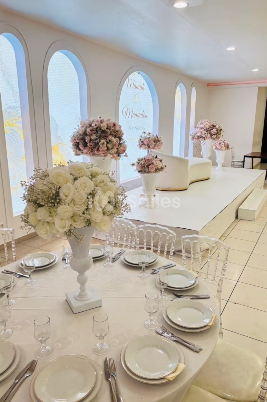 Salle de réception décorée avec des fleurs blanches et roses, table dressée et estrade avec canapé en arrière-plan.