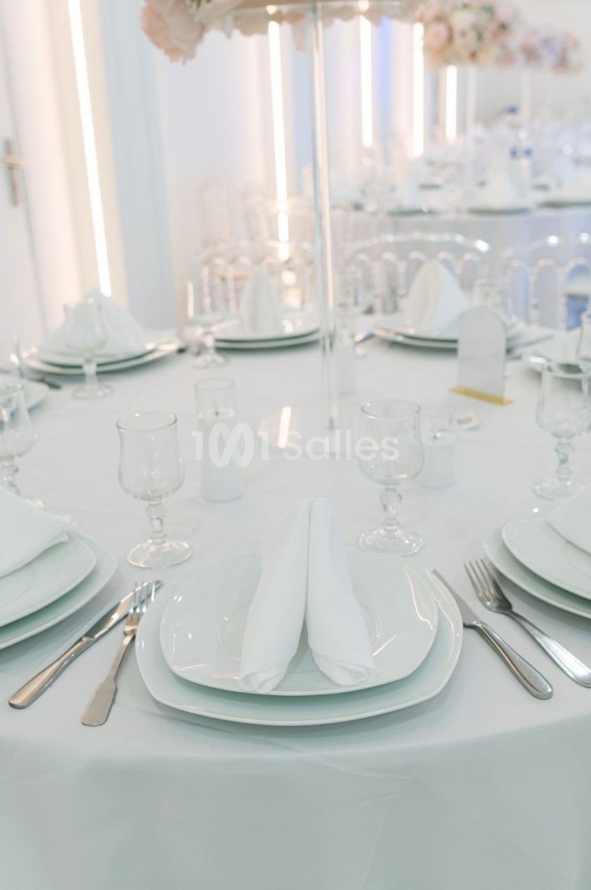 Table dressée avec nappes blanches, assiettes, couverts, verres et serviettes pliées dans un décor lumineux et élégant.