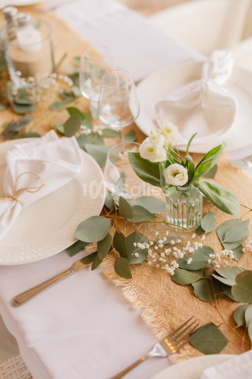 Table dressée avec assiettes blanches, verres et petits vases de fleurs blanches sur chemin de table en jute.