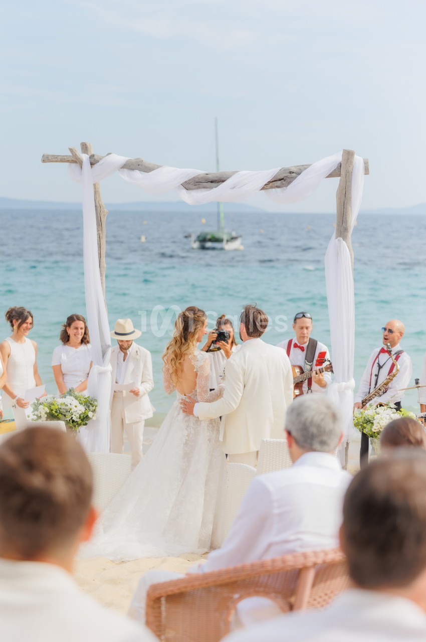 Cérémonie de mariage sur la plage avec arche drapée blanche face à la mer.