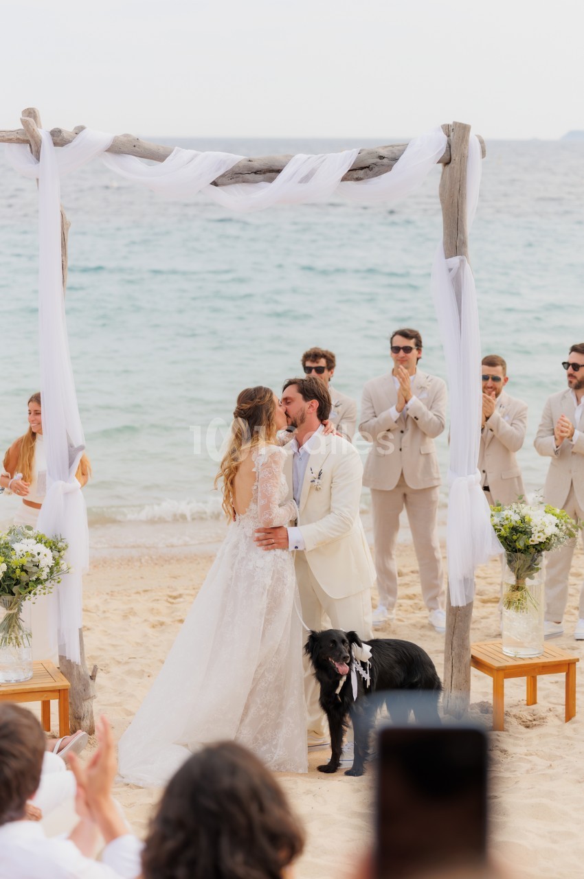 Cérémonie de mariage sur la plage avec arche en bois et voilage blanc, mariés face à face et invités debout autour.