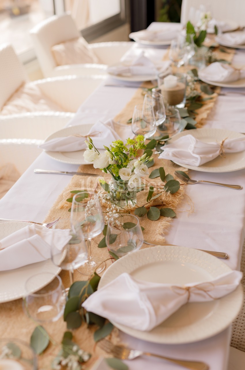 Table dressée avec assiettes blanches, serviettes beiges et compositions florales blanches le long du chemin de table.
