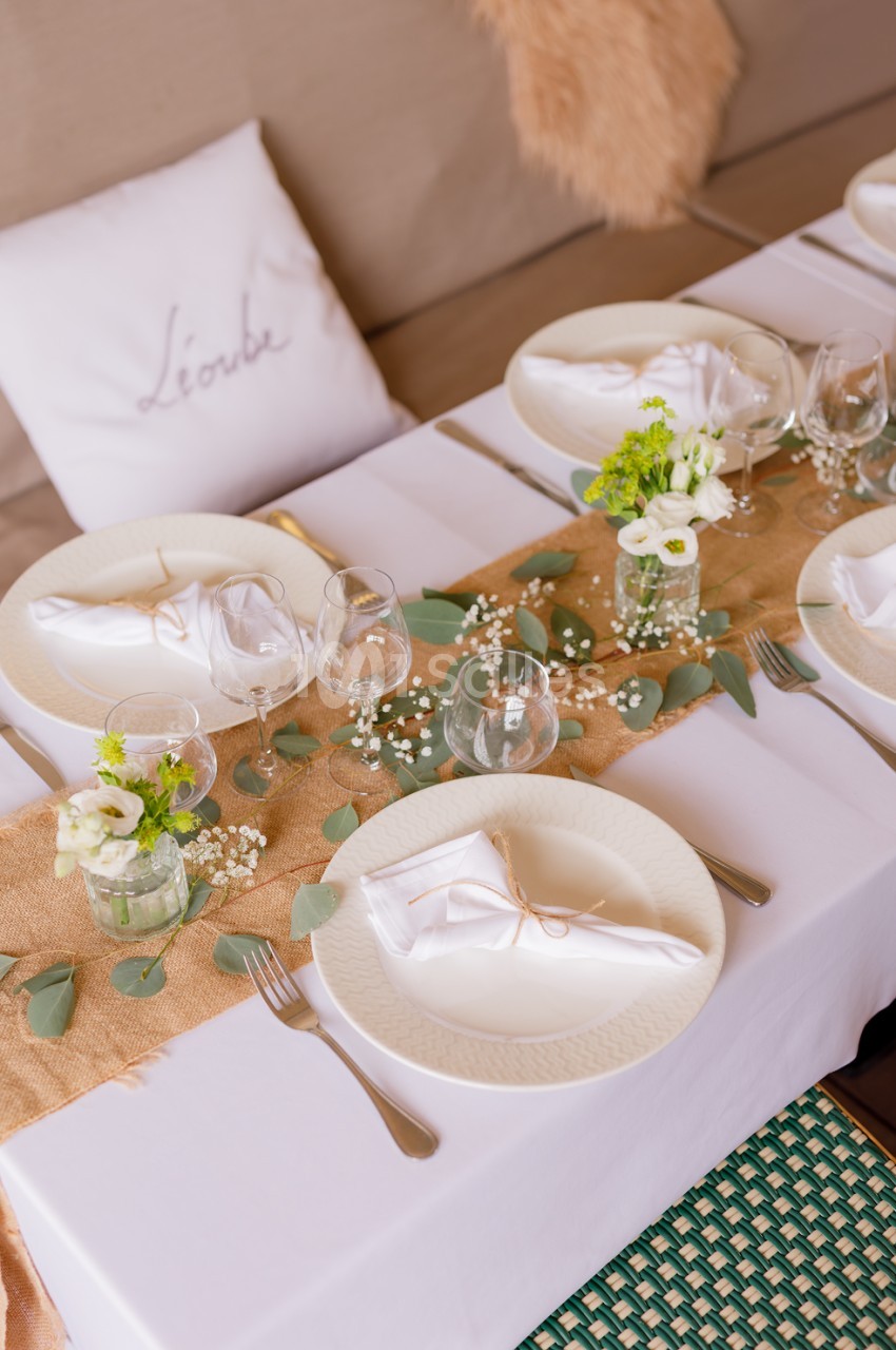 Table dressée avec nappes blanches, chemin de table jute, assiettes, verres et petits bouquets blancs et verts.