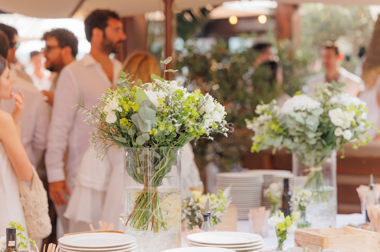 Table dressée avec bouquets blancs et jaunes en vases, convives debout en arrière-plan sous voile.