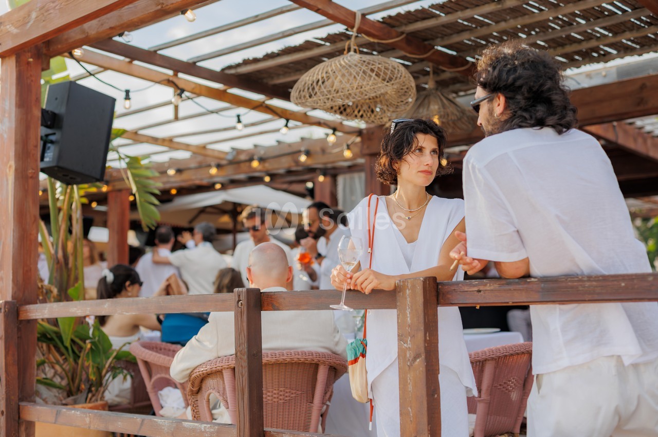 Terrasse couverte en bois avec poutres apparentes, luminaires suspendus et convives attablés lors d'un événement.