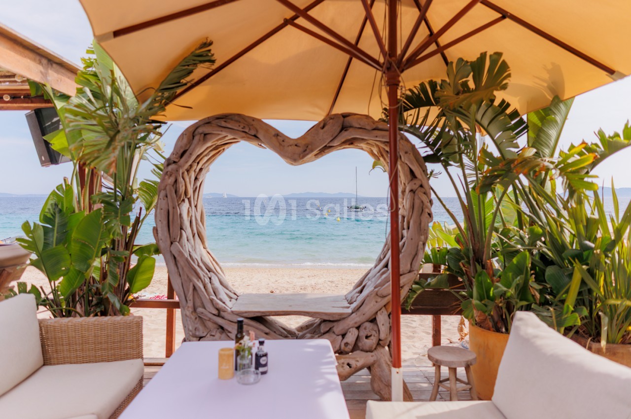 Terrasse de plage avec auvent beige, structure en bois en forme de cœur, plantes tropicales, table blanche et vue sur la mer.