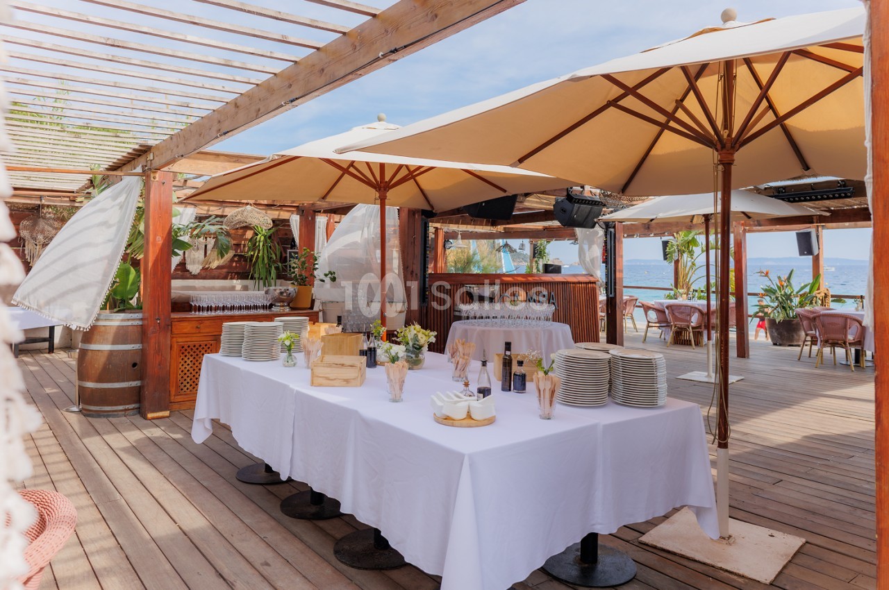 Terrasse en bois avec table dressée sous parasols beige, vue mer en arrière-plan.