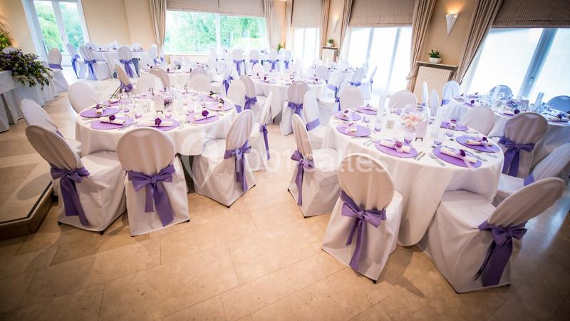 Salle de réception décorée avec des tables rondes, nappes blanches et chaises ornées de nœuds violets.